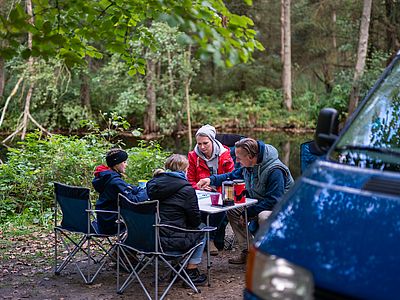 Eine Familie sitzt beim Camping an einem Tisch im Wald und spielt gemeinsam. Auf